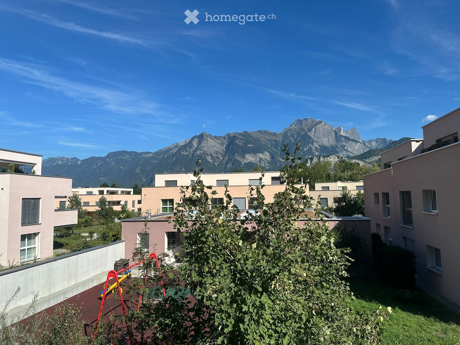 Residential complex with modern buildings and a playground, surrounded by greenery and mountains in the background.