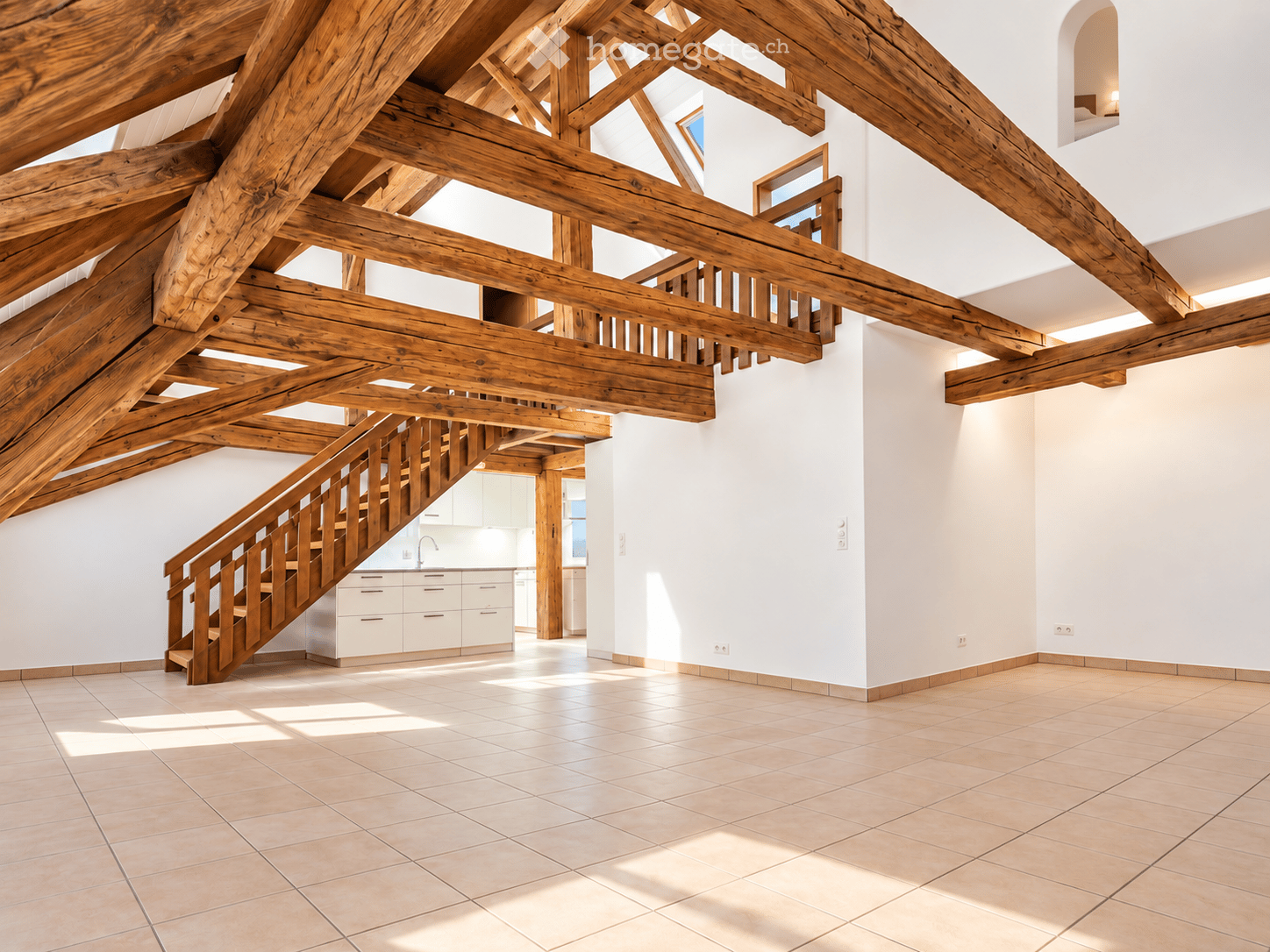 Spacious room with exposed wooden beams, a wooden staircase leading to an upper level, and a small kitchen area with white cabinets and a sink.
