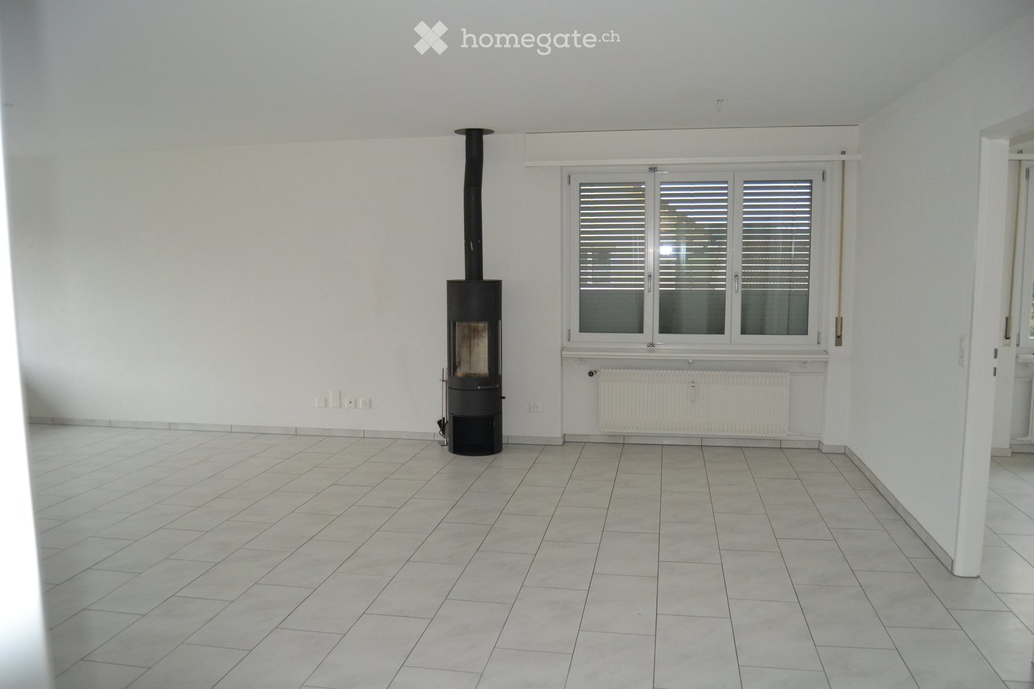 Empty room with tiled floor, white walls, large window with blinds, and a black wood-burning stove with chimney.