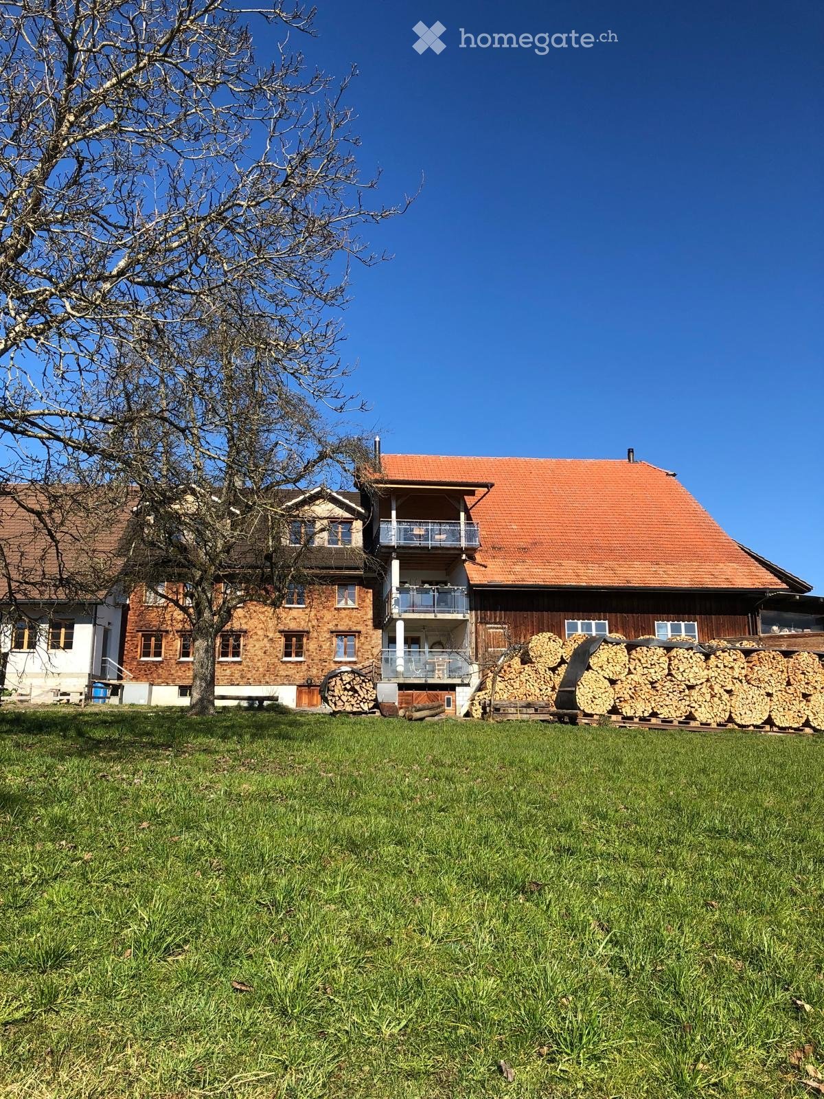 Two-story house with red-tiled roof, brick and wooden exterior, balcony, and stacks of firewood in front.