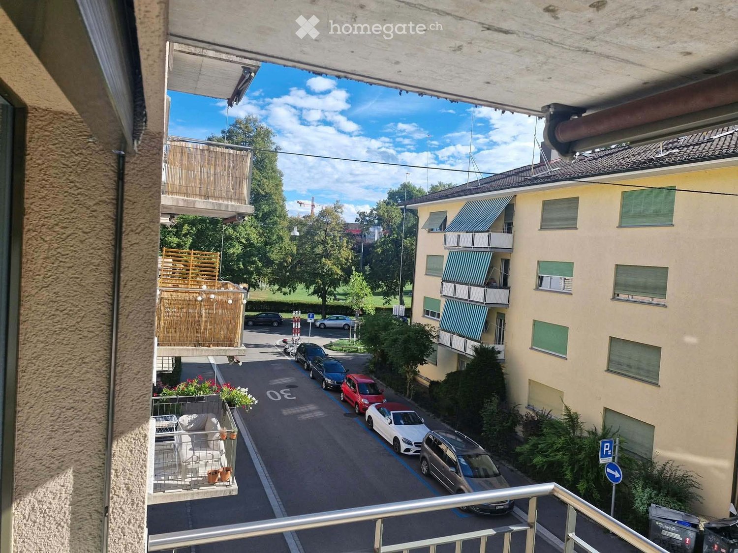 Balcony overlooking a street with parked cars, buildings, and greenery. Features a metal railing, a small table, chairs, and flower pots.
