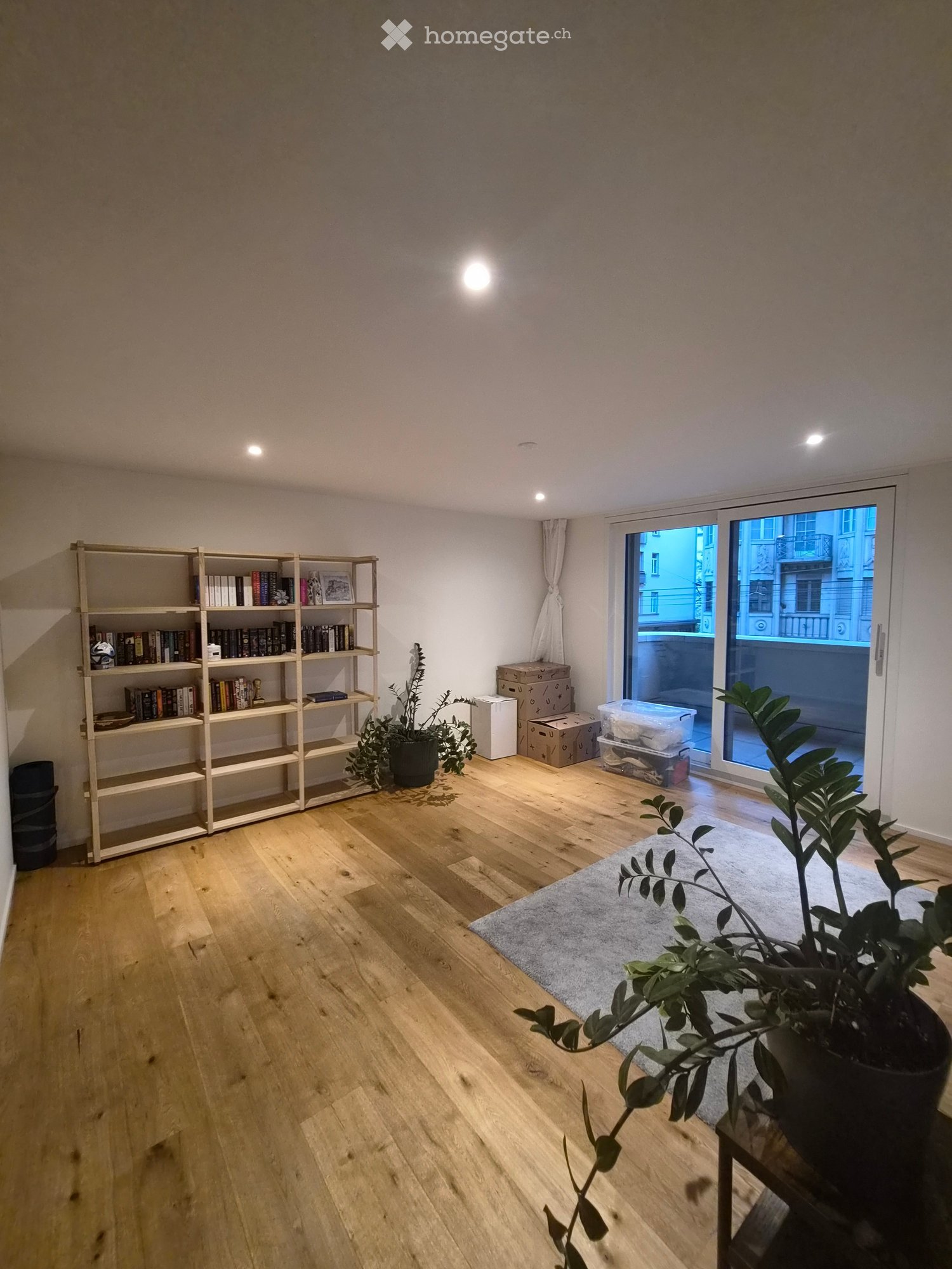 Bright, minimalist room with wooden flooring, large window with view, bookshelf filled with books, potted plants, and storage boxes near the window.