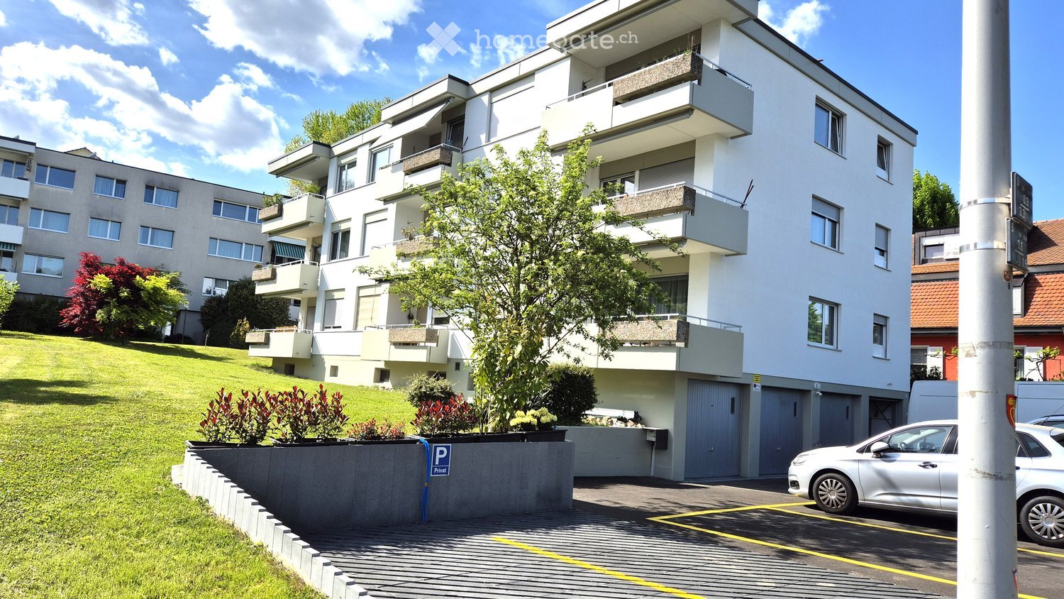Modern multi-story apartment building with balconies, surrounded by greenery and a parking area.