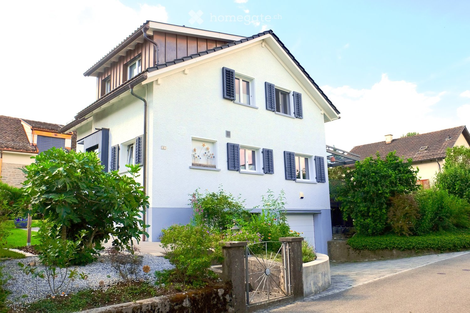 Two-story house with white exterior, dark shutters, and a garage. The house features a well-maintained garden with various plants and a decorative metal gate.