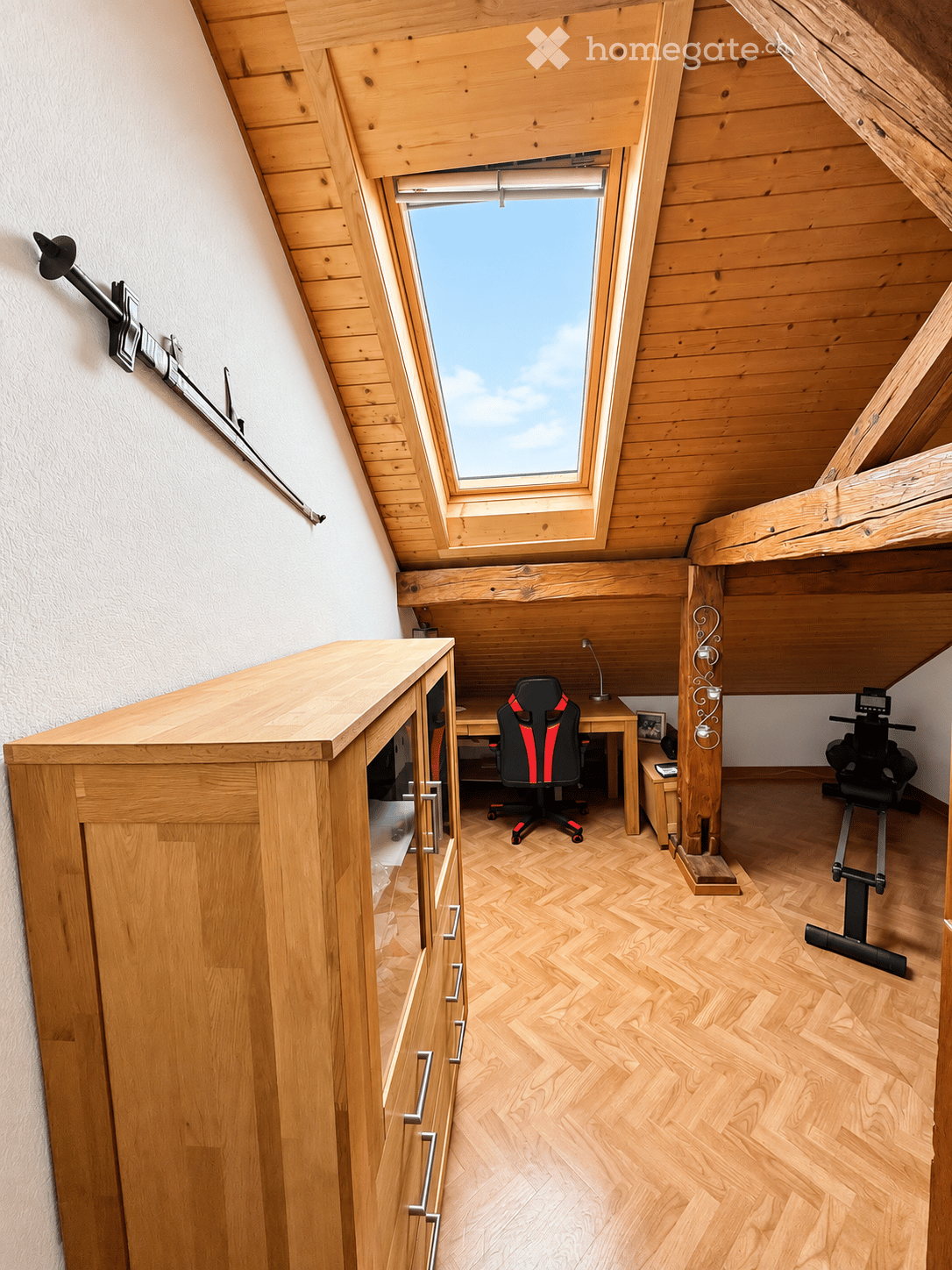 A well-lit attic room with wooden walls and herringbone flooring, featuring a desk with a chair, a cabinet, and an exercise bike.