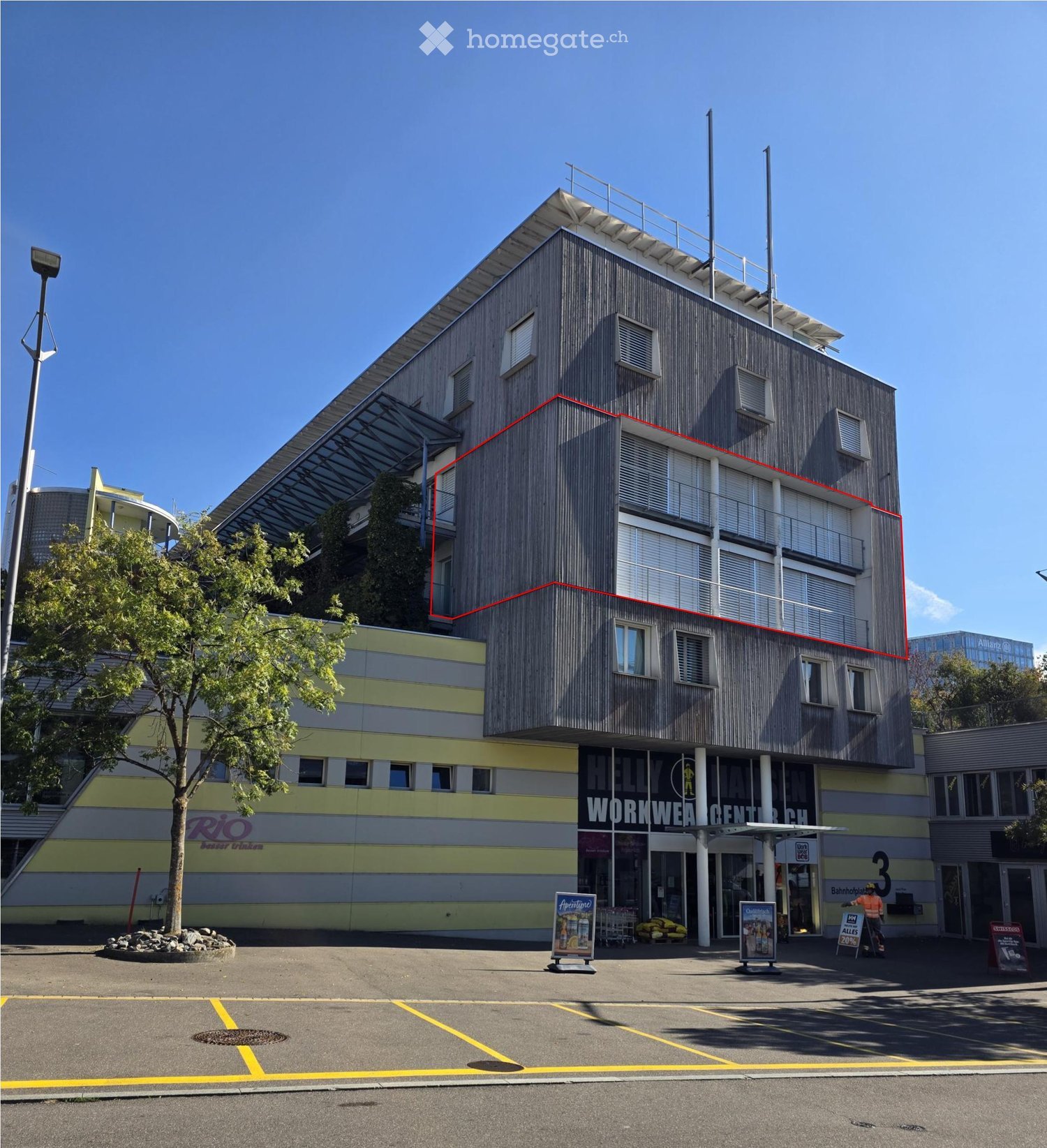 Modern multi-story building with a combination of grey and yellow facades, featuring large windows, a raised ground floor, and a parking area.
