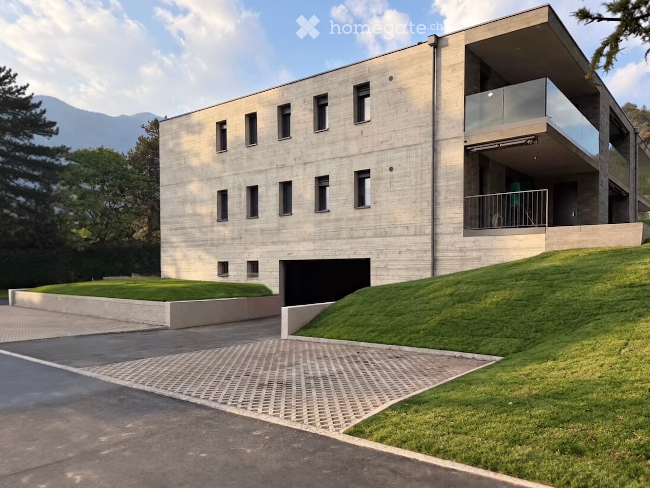 Modern concrete house with multiple windows, raised ground floor, garage, and a balcony on the upper level.