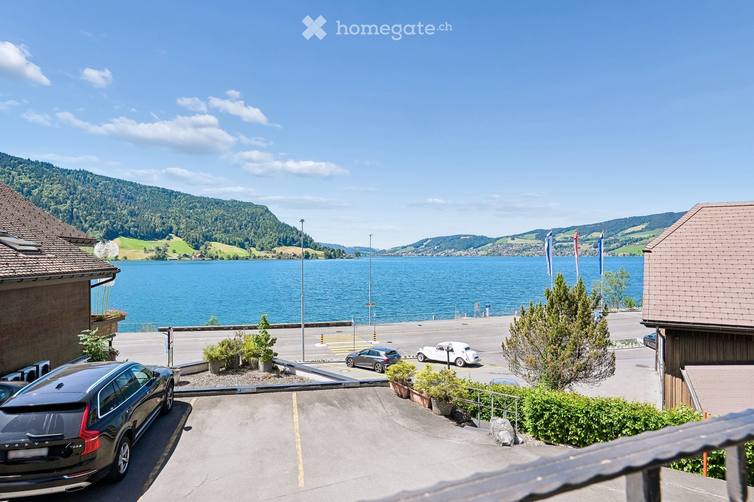 View from a balcony overlooking a parking area, lake, and surrounding hills with flags and parked cars.