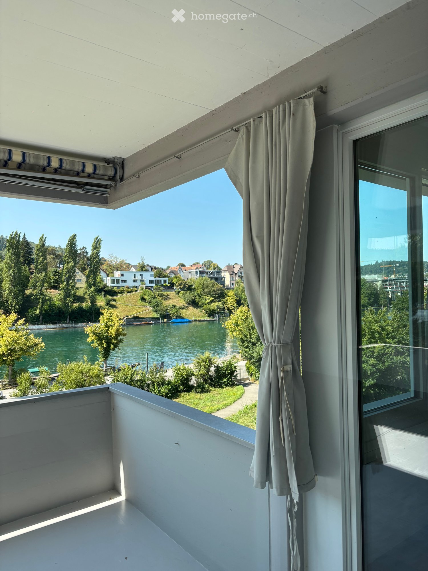 Balcony with white railing, beige curtains, and a scenic view of a river and surrounding greenery.