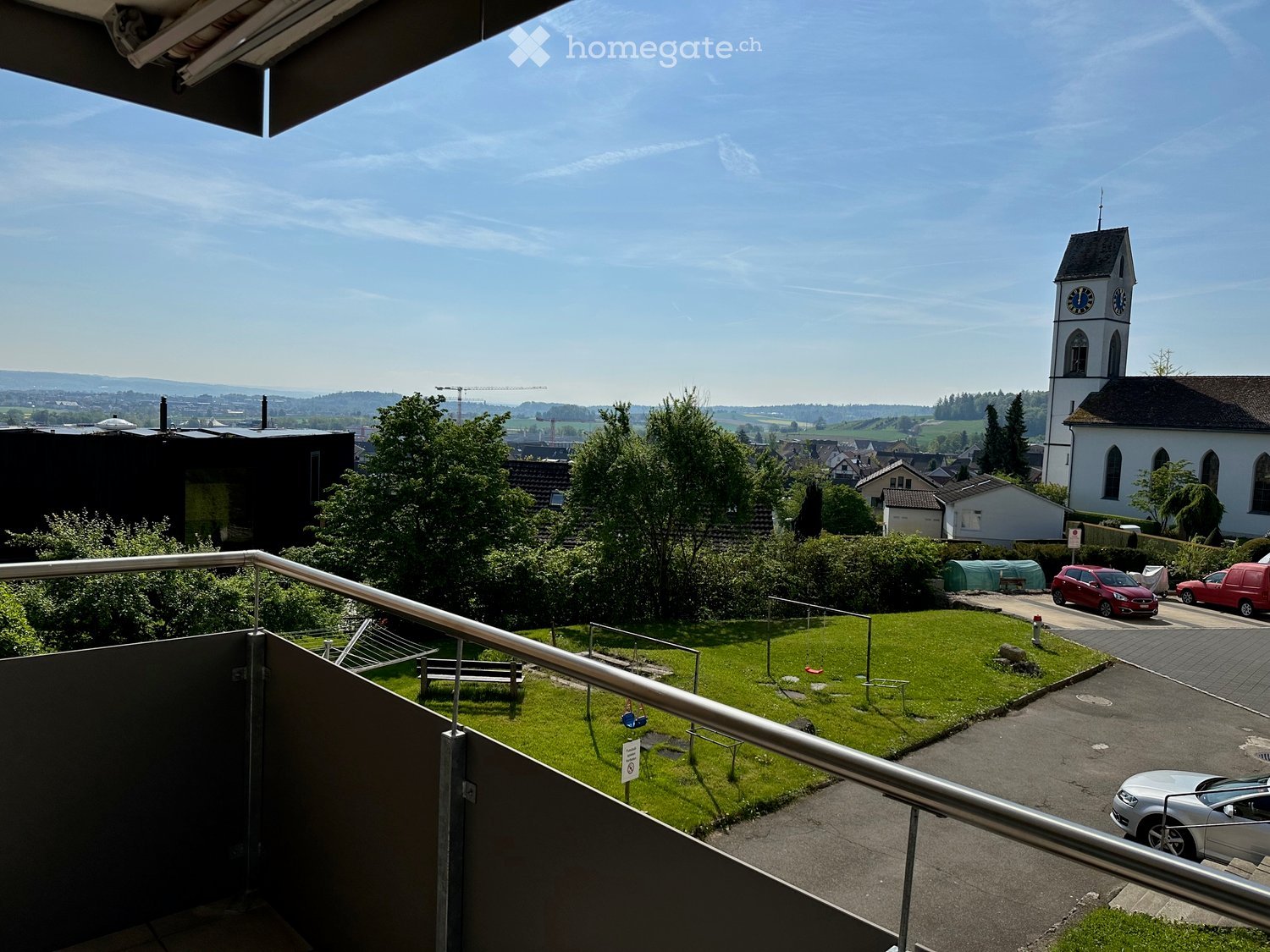 Balcony overlooking a green lawn, playground, and a town with a church tower in the background.