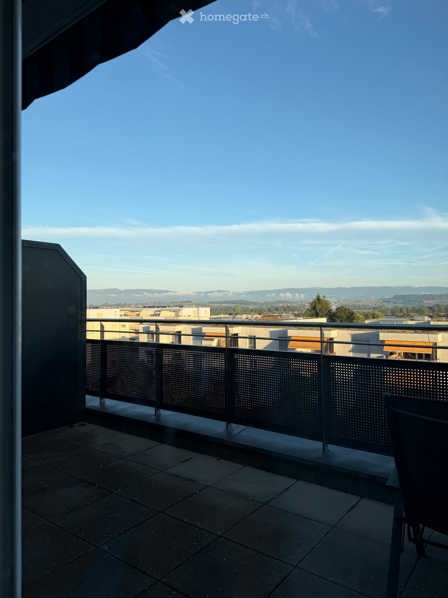Balcony with a metal railing, tiled floor, and a scenic view of a cityscape and distant hills under a clear blue sky.