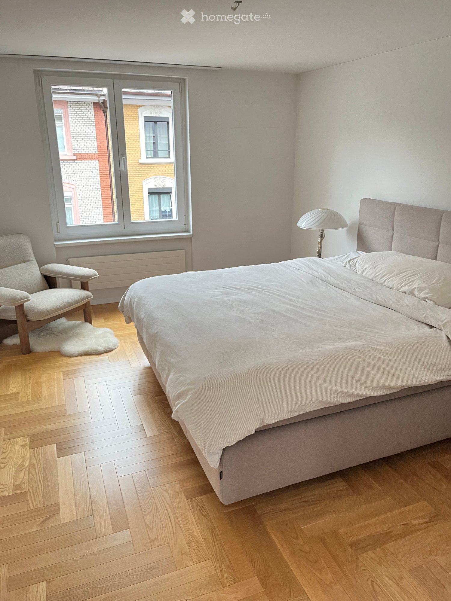 Bright bedroom with wooden flooring, large window, bed with white bedding, armchair, and a lamp on the nightstand.