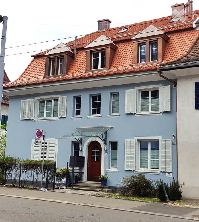 Two-story house with light blue exterior, red-tiled roof, white shutters, and a central entrance with a small awning.