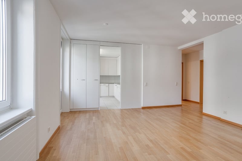 Empty room with light wooden flooring, white walls, large built-in closet, and a view into an adjacent kitchen area.