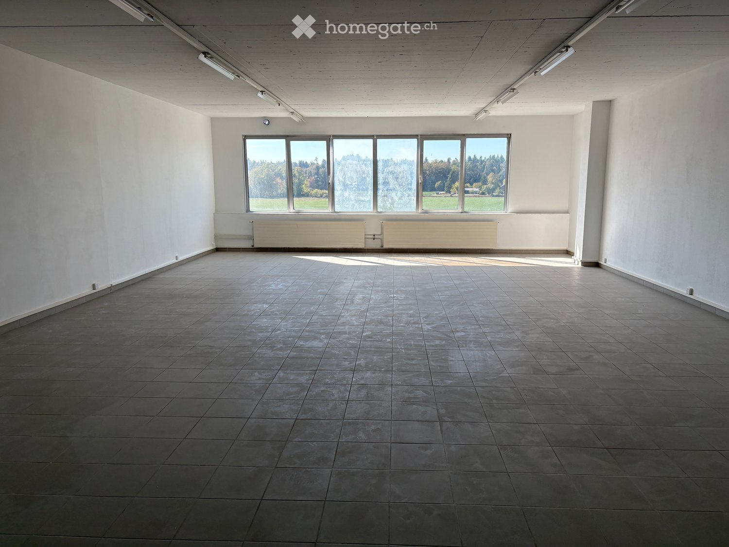 Empty room with tiled floor, white walls, large windows, and a radiator under the windows.