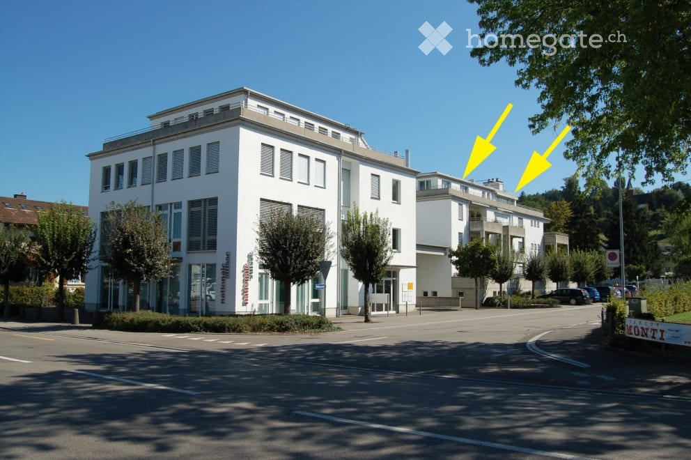 Modern multi-story building with white facade, large windows, and a flat roof, surrounded by trees and parked cars.