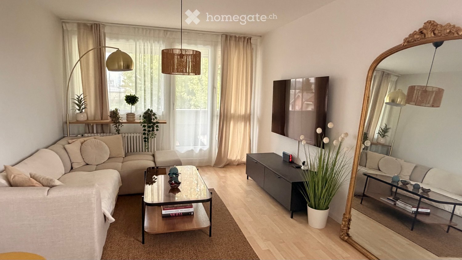 Modern living room with a beige sofa, glass coffee table, large mirror, floor lamp, and decorative plants.