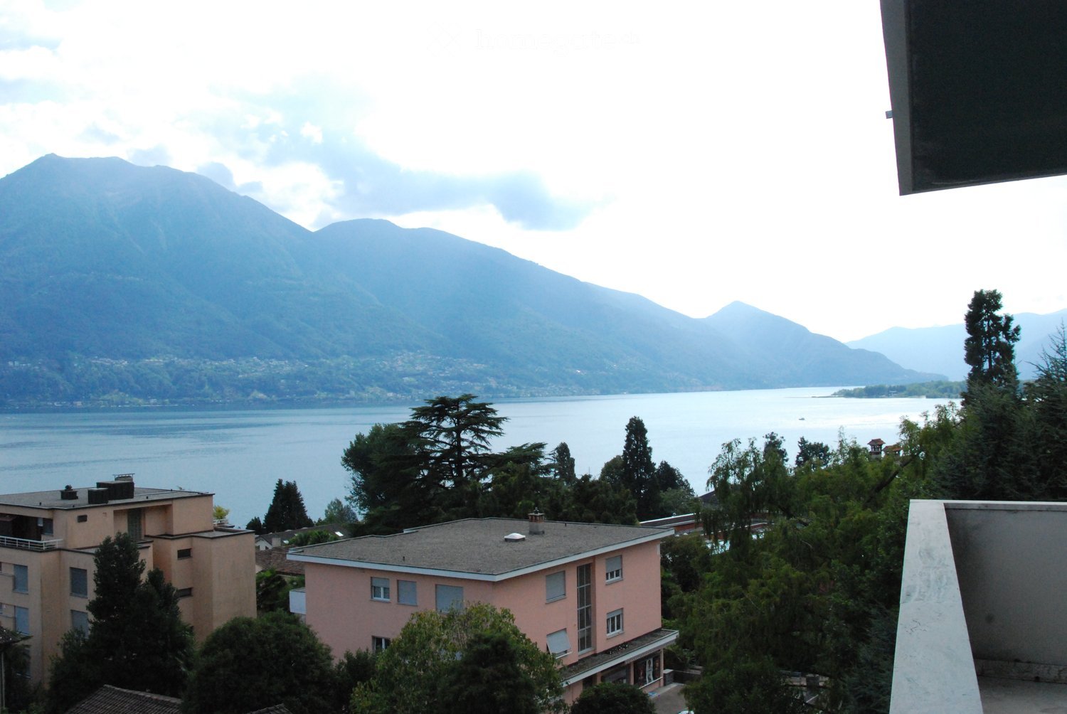 View from a balcony showing a lake, mountains, and surrounding buildings with trees.