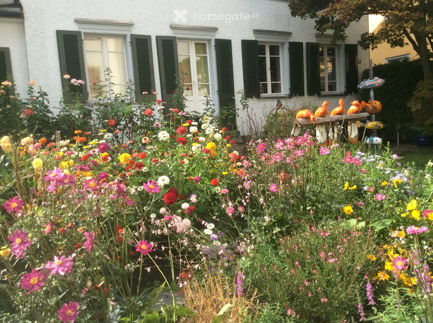 A colorful garden with a variety of flowers, a white house with green shutters in the background, and decorative bird sculptures on a wooden bench.