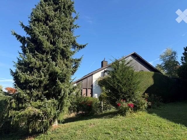 Single-family house with a pitched roof, white exterior, and dark wooden siding on one side. The house is surrounded by a well-maintained lawn, tall evergreen trees, and flowering shrubs.
