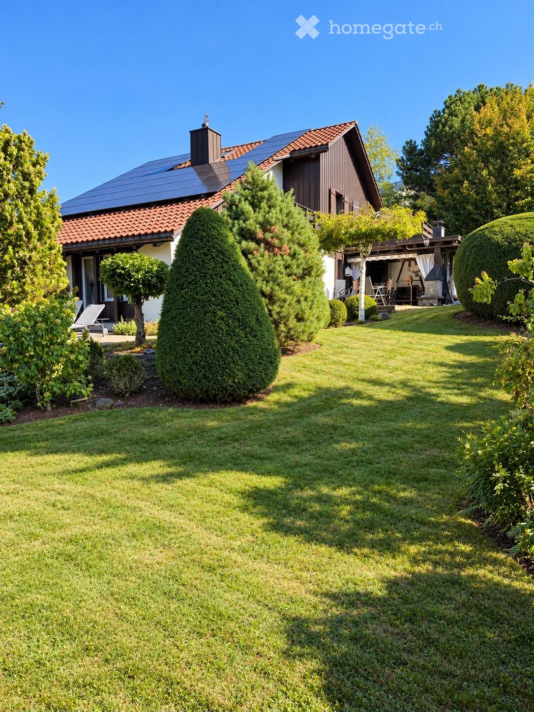Well-maintained garden with a neatly trimmed lawn, various trees and shrubs, and a patio area with outdoor furniture near the house.
