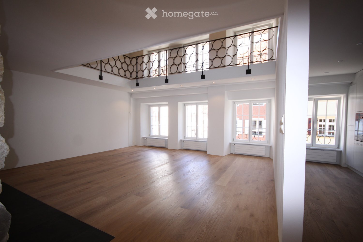 Large empty room with wooden floor, white walls, three windows with radiators, black railing on raised ground floor