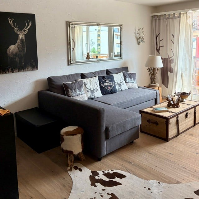 Modern living room with grey sectional sofa, decorative pillows, coffee table, floor lamp, and a large mirror above the sofa. The room features wooden flooring, a cowhide rug, and a deer head mounted on the wall.