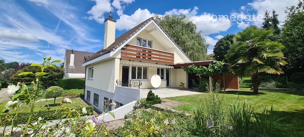 Two-story house with a modern design, featuring a balcony, large windows, and a well-maintained garden with various plants and trees.