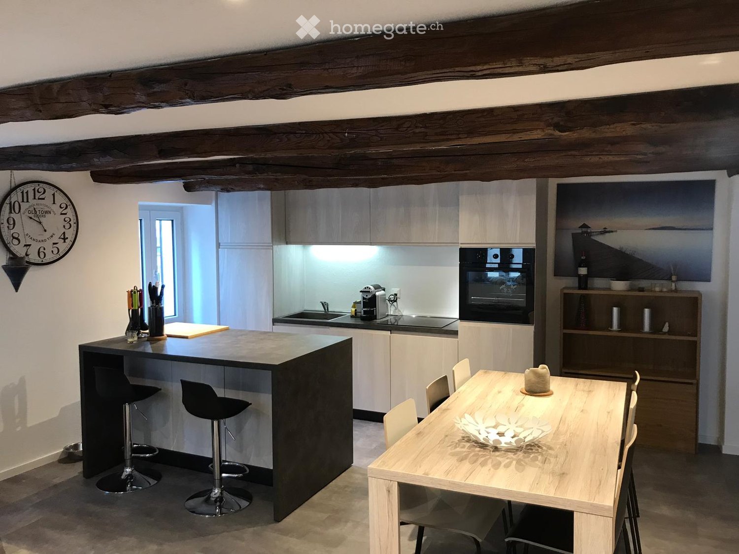 Modern kitchen with a central island, bar stools, dining table, and chairs. Features include a clock on the wall, an oven, sink, and various kitchen appliances.