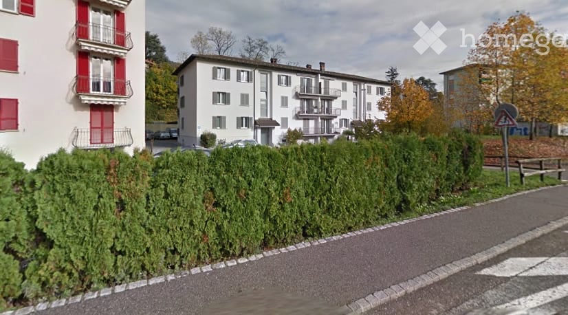 Multi-story residential building with balconies, surrounded by greenery and a paved road.