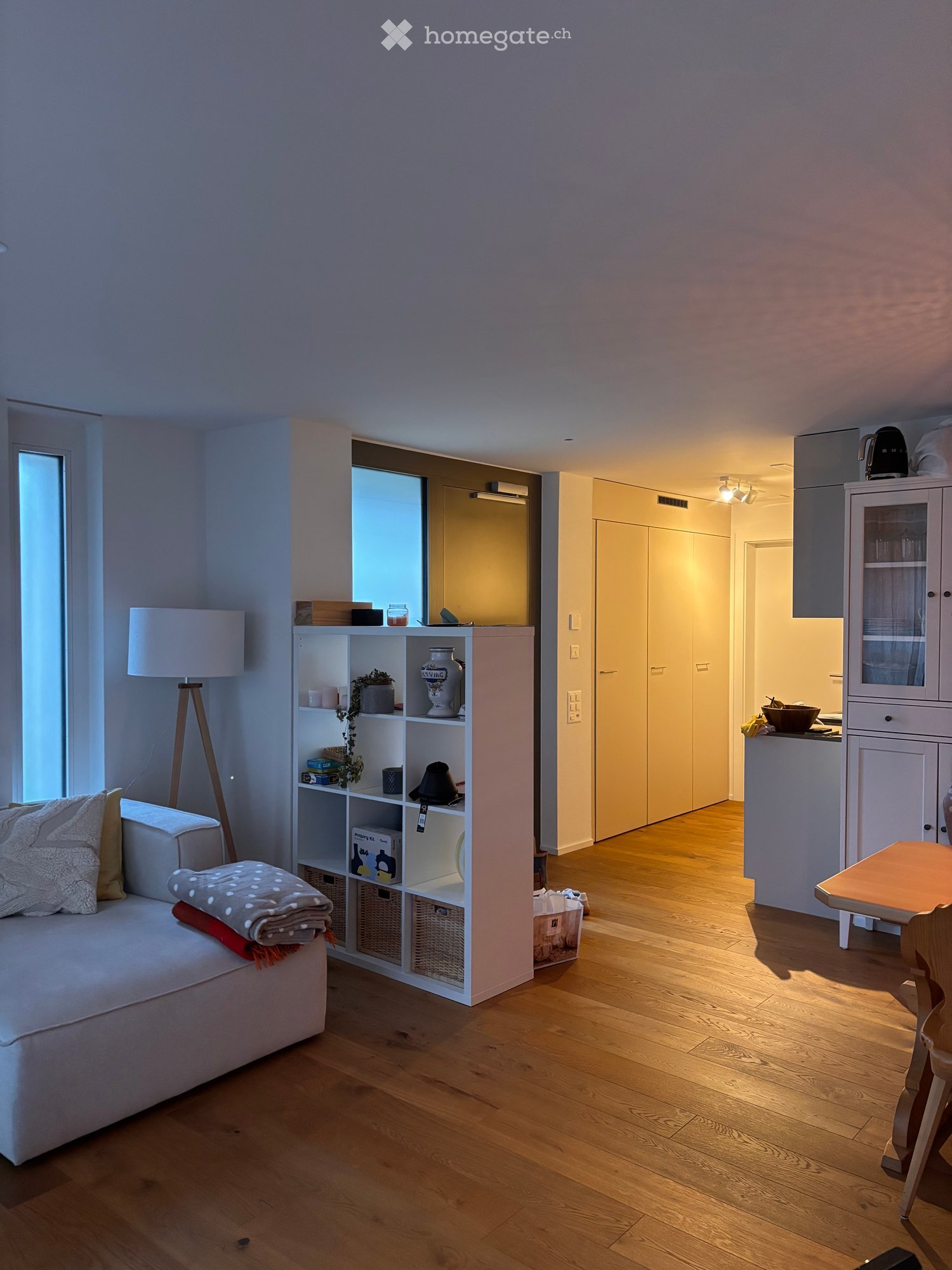 A modern, well-lit living room with a white sofa, a wooden table, a shelf with decorative items, and a lamp. The room features light wooden flooring and large windows allowing natural light to fill the space.