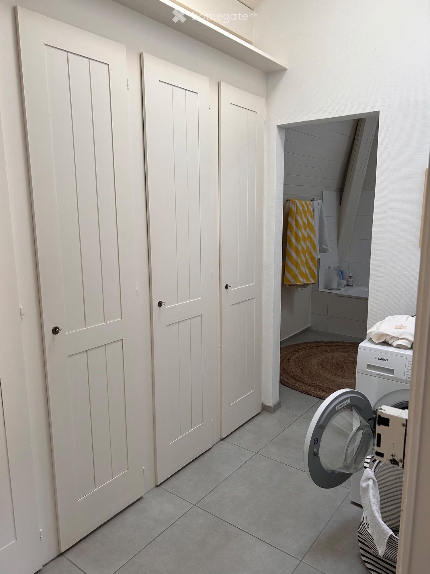 A small bathroom with a washing machine, a sink, and a towel hanging on the wall. The room has white doors and tiled flooring.