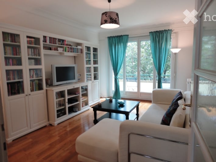 Bright living room with large windows and a view, featuring a white sectional sofa, a black coffee table, a television on a white cabinet, built-in bookshelves, and a floor lamp.