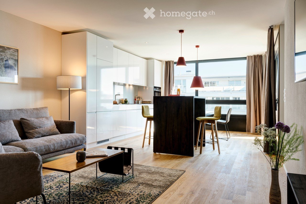 Modern living room with a grey sofa, coffee table, wooden floor, pendant lamps, and a small kitchen area with white cabinets and a bar counter.