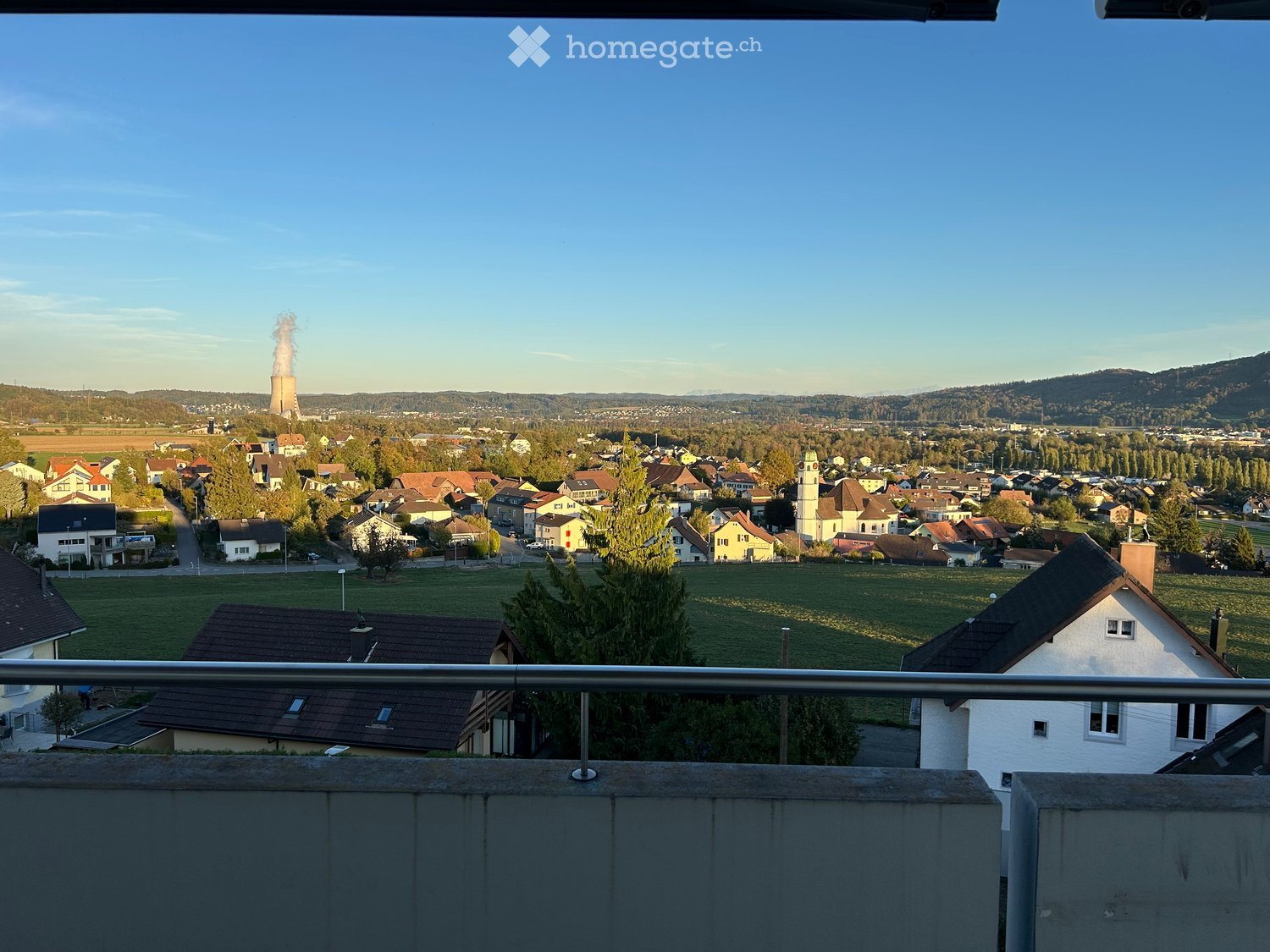 Balcony overlooking a town with houses, green fields, and a distant industrial plant emitting smoke.