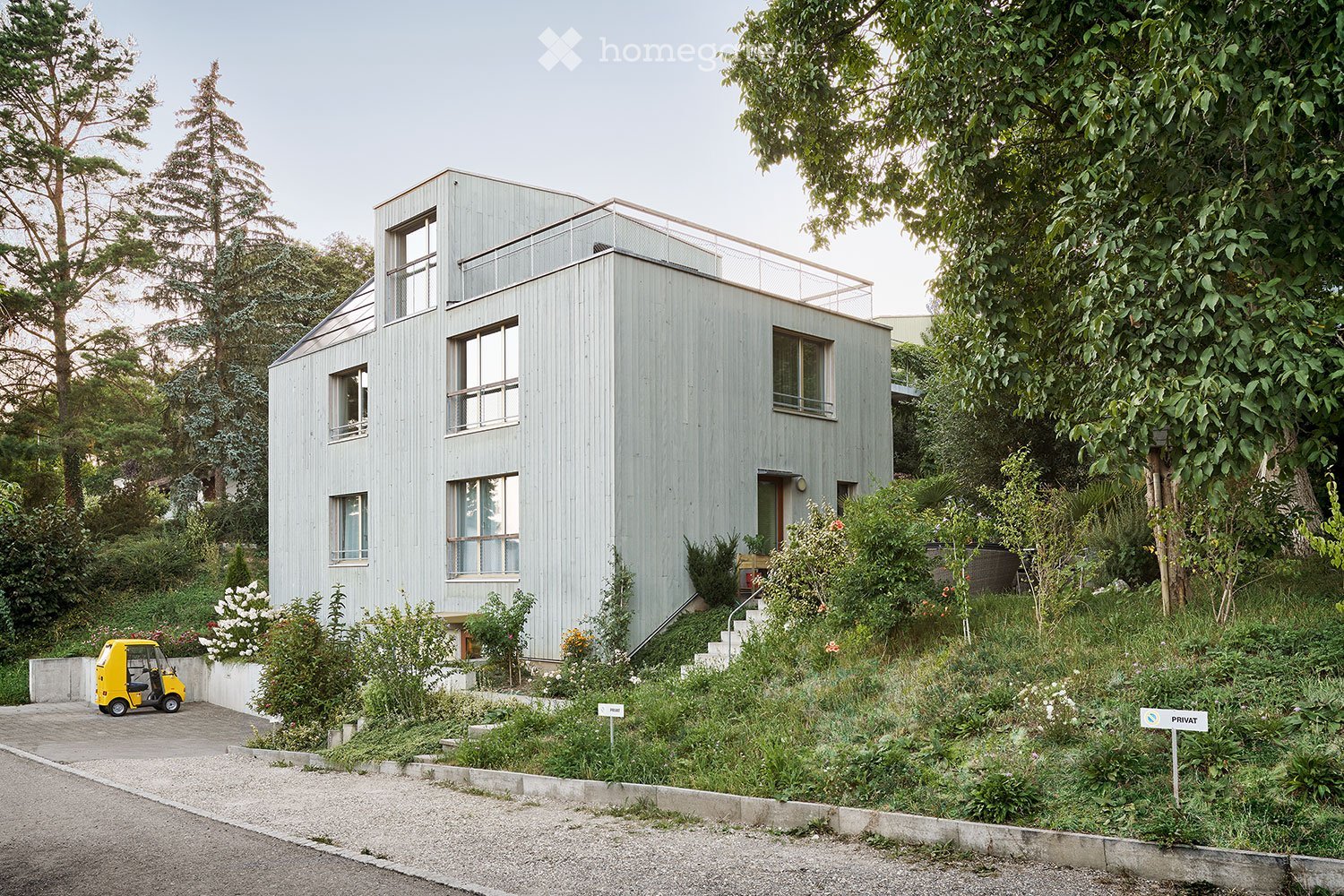 Modern two-story house with light grey wooden exterior, large windows, and a raised ground floor surrounded by lush greenery and trees.