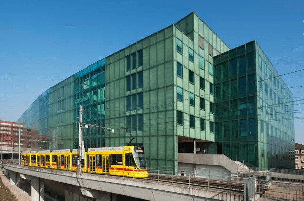Modern green glass building with a yellow and red tram passing by on an elevated track.