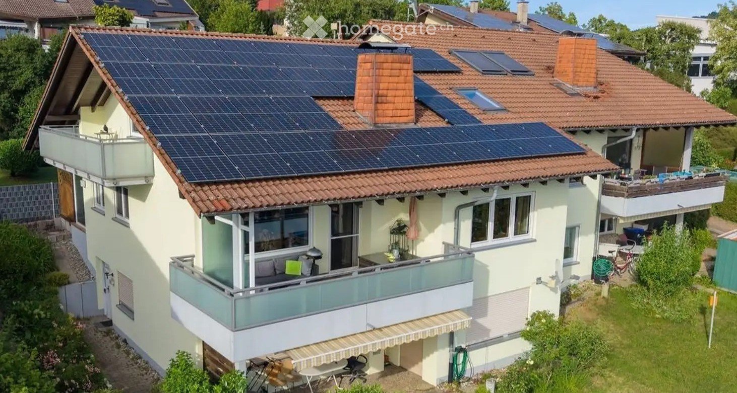 Two-story house with solar panels on the roof, multiple balconies, and surrounding greenery.