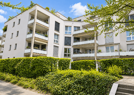 Modern multi-story apartment building with white facade, multiple balconies, and greenery in front.