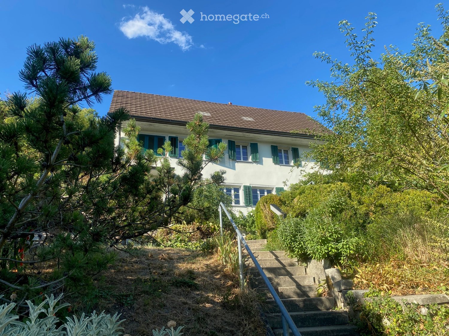 Two-story white house with green shutters and a brown tiled roof, surrounded by trees and shrubs. A concrete staircase leads up to the house.