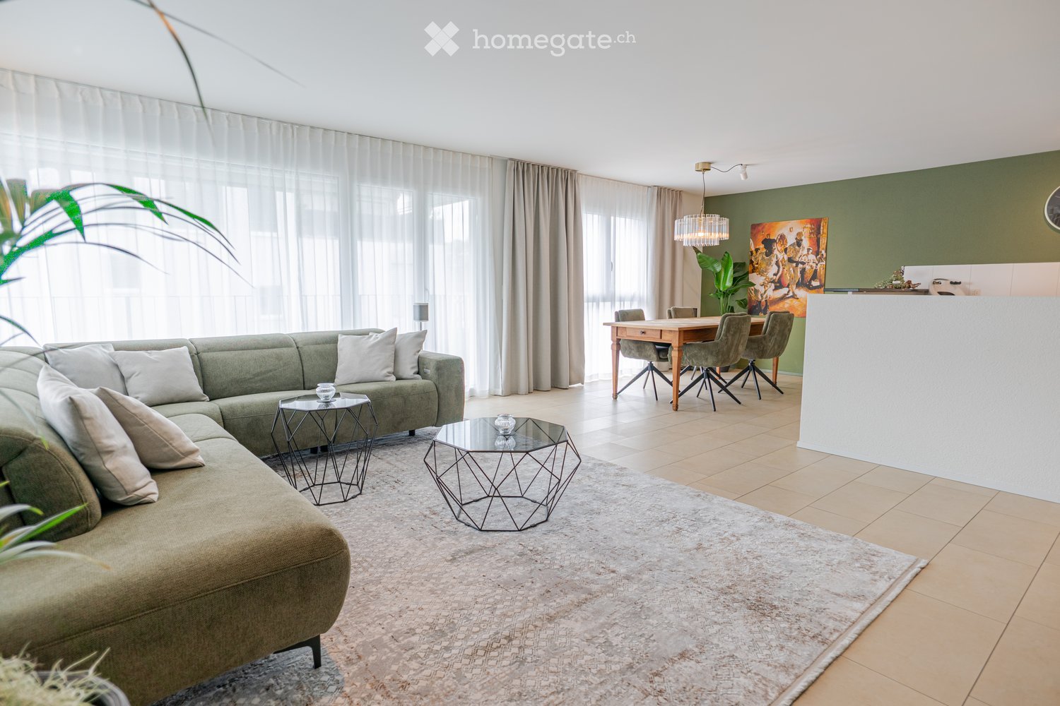 Modern living and dining area with a green sectional sofa, glass coffee tables, a dining table with chairs, and a chandelier.