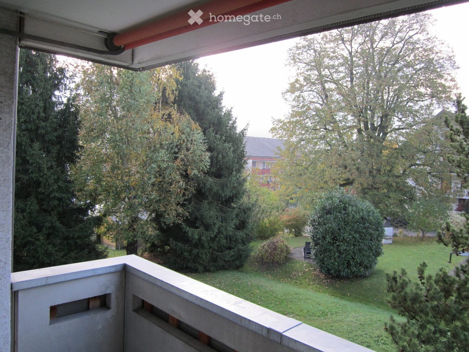 Balcony overlooking a well-maintained garden with various trees and shrubs, featuring a concrete railing and a covered section.