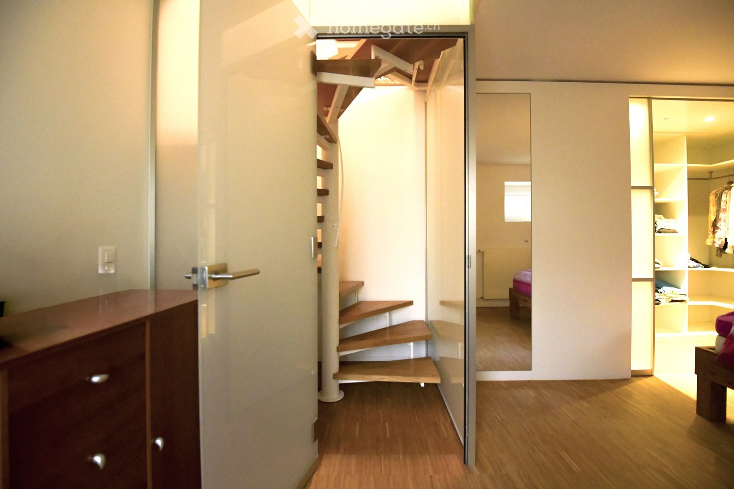 A narrow hallway with wooden flooring, a small staircase leading upwards, and an open doorway to a bedroom area with a bed and wardrobe visible.