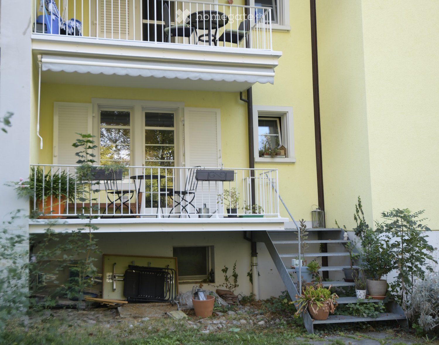 Balcony with a table, chairs, and potted plants, attached to a yellow building with multiple windows and an awning.