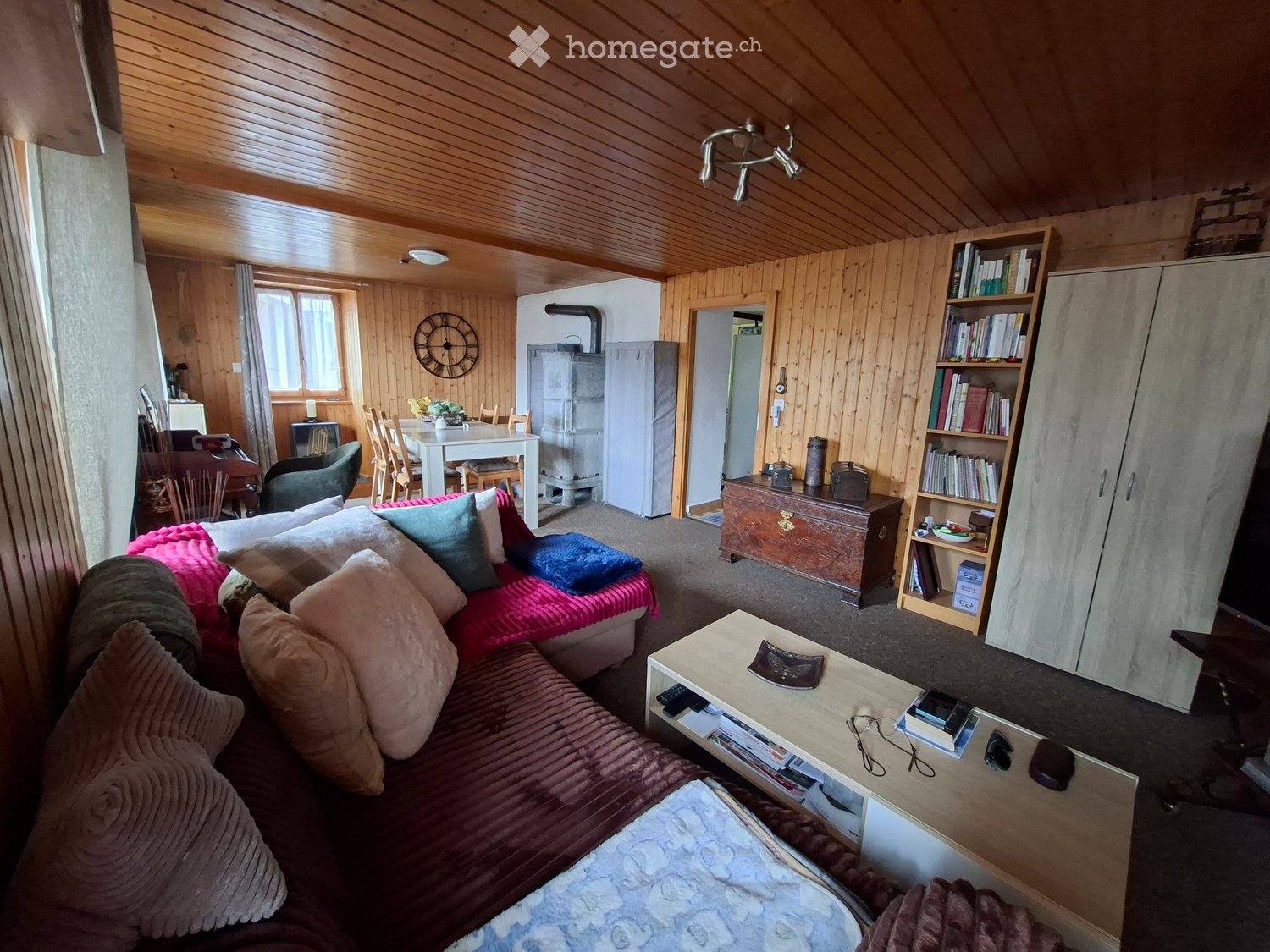 Cozy living room with wooden walls and ceiling, featuring a red sofa, coffee table, bookshelf, dining area with table and chairs, and a refrigerator in the background.