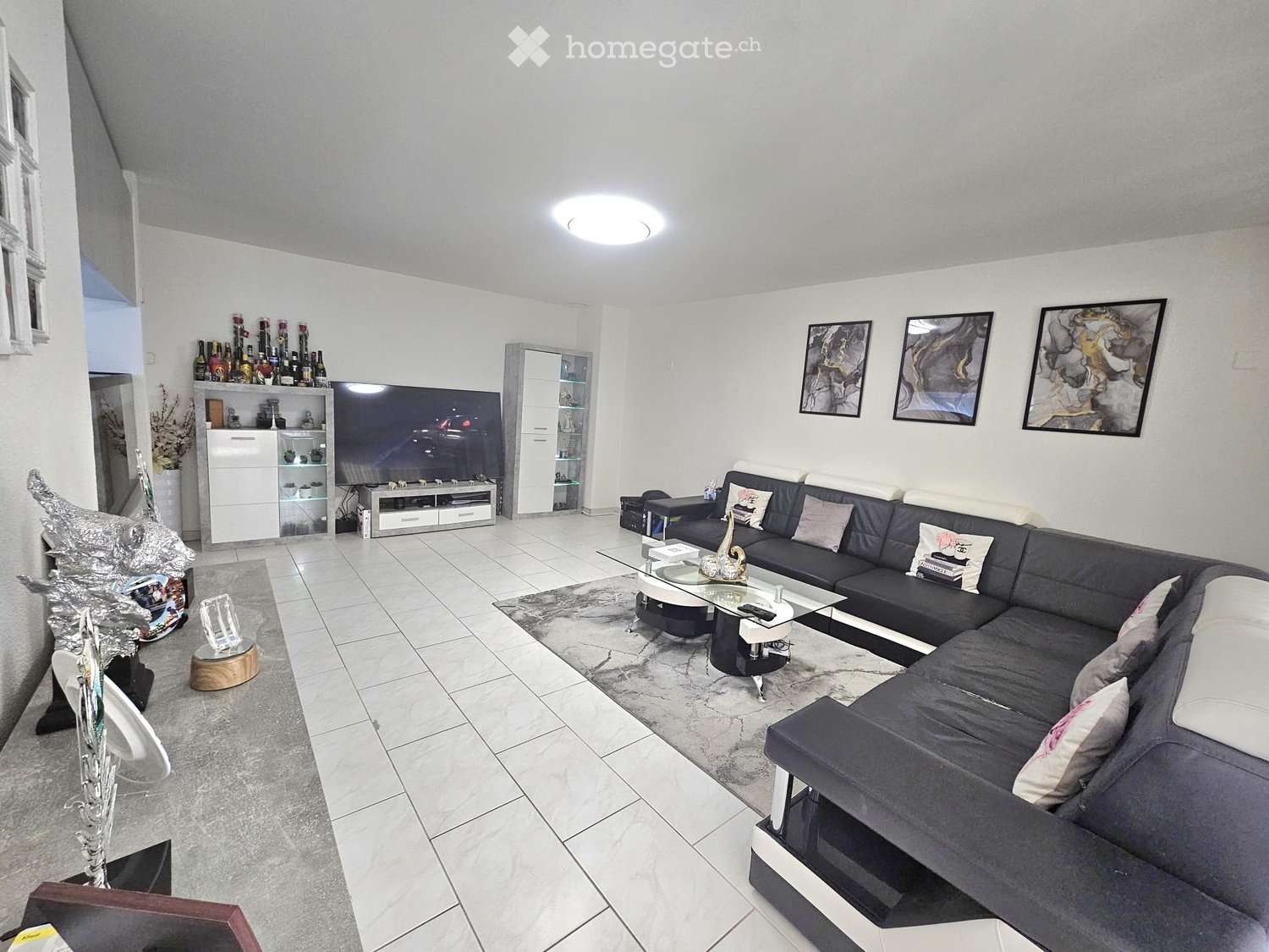 Modern living room with a large sectional sofa, coffee table, TV stand, and decorative shelves. The space features white walls, tiled flooring, and contemporary decor including artwork and a vase.