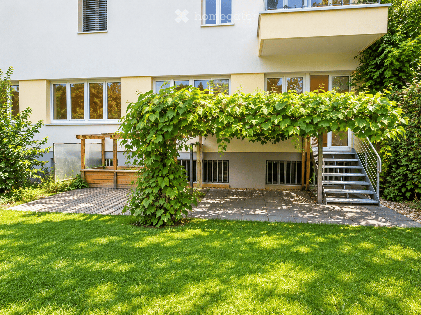 Well-maintained garden with green grass, a wooden pergola, a small staircase leading to a raised terrace, and a climbing plant providing shade.