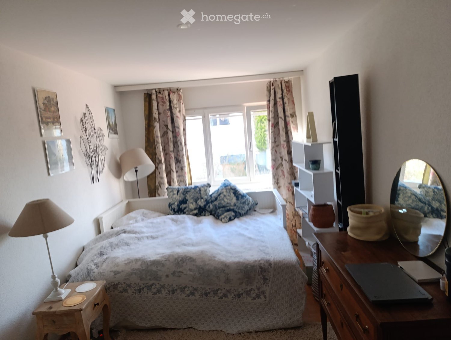 Cozy bedroom with a double bed, nightstand, lamp, mirror, laptop, and decorative items including a vase and basket. The room features large windows with curtains and artwork on the walls.