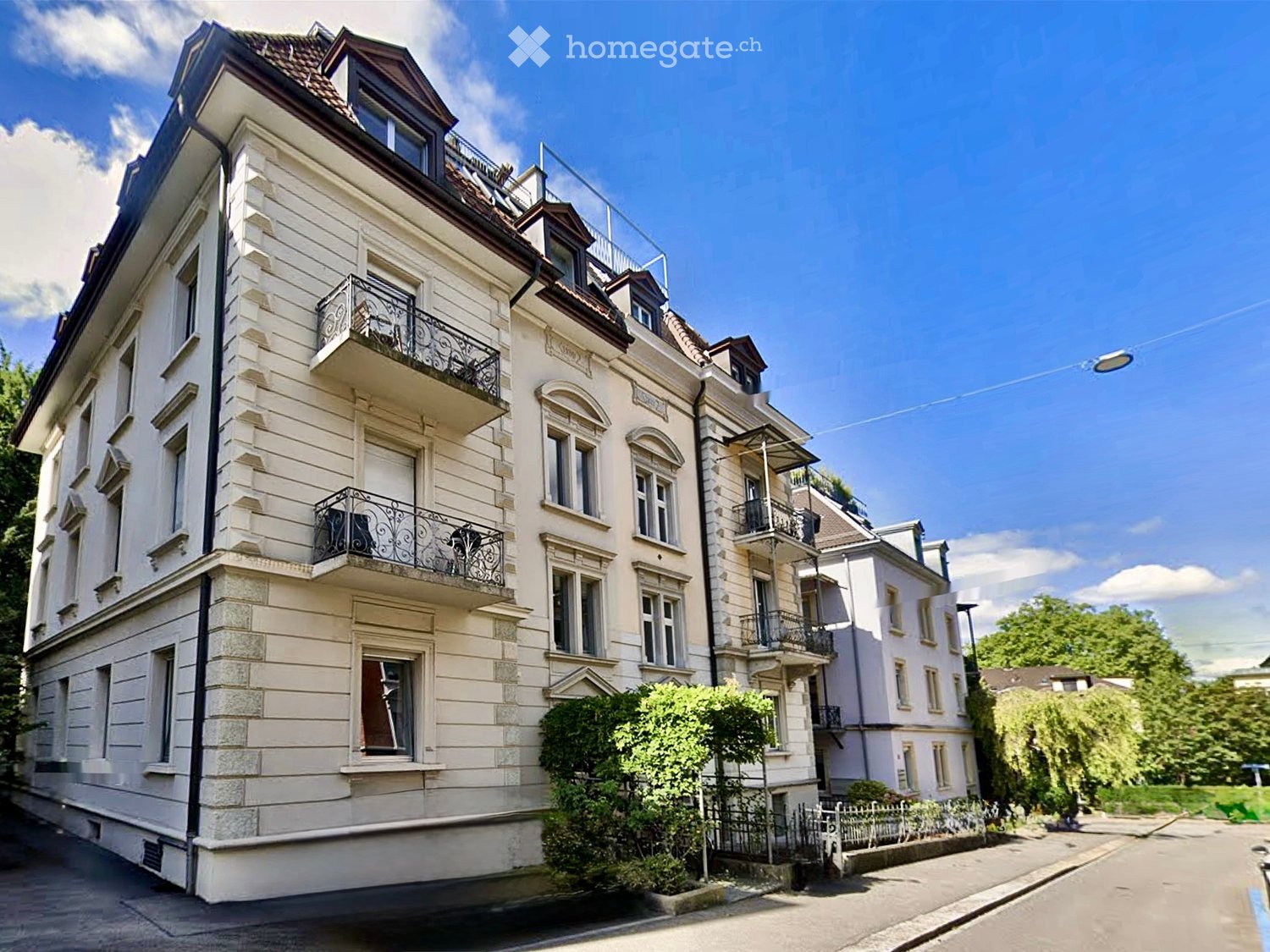 A multi-story residential building with light-colored facade, multiple balconies with ornate railings, and a well-maintained garden in front.