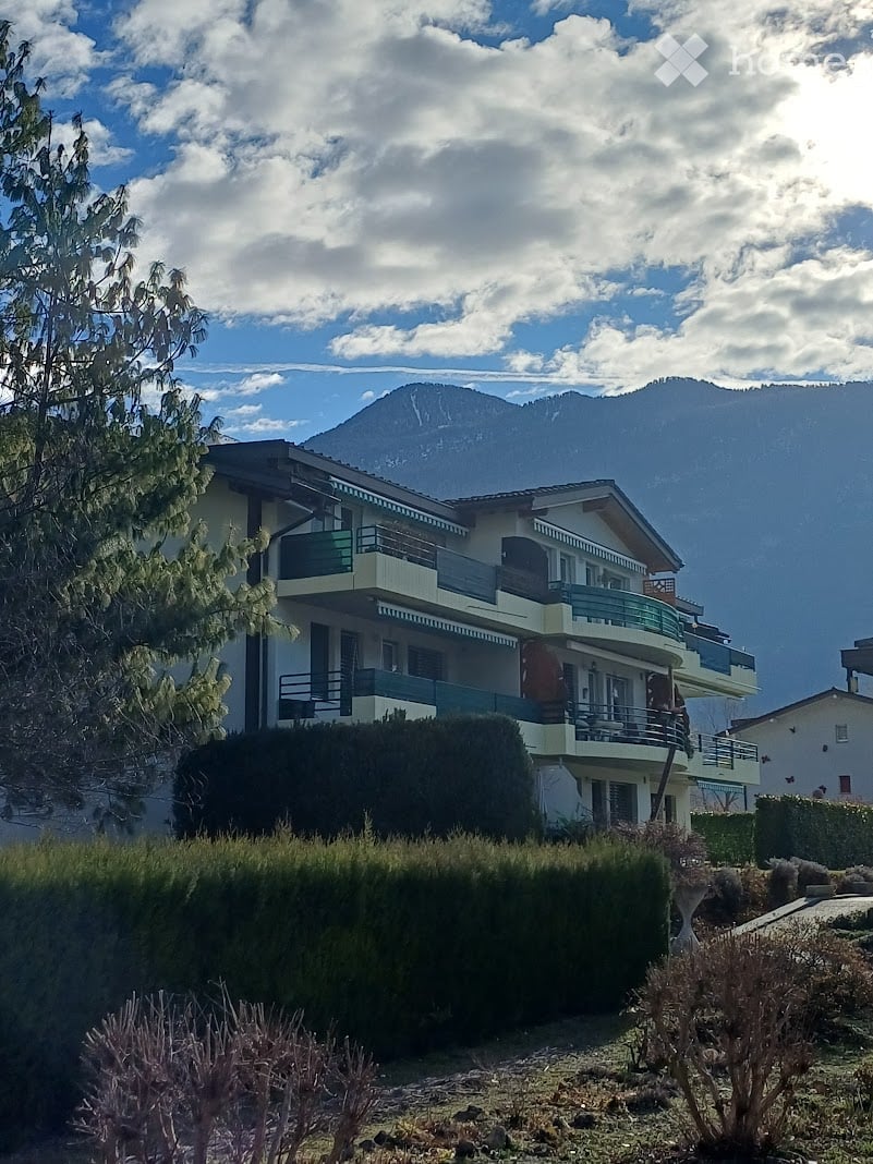 Two-story house with balconies, surrounded by greenery and a mountain view.